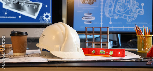 White hard hat and blueprints on an engineer's desk with computer screens displaying technical drawings and design plans