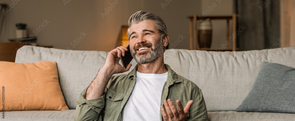 Fototapeta premium Happy Senior Man Smiling While Talking On Smartphone In Modern Living Room, Engaged In Conversation At Home. Cheerful Mature Caucasian Male Enjoying Interaction In Comfortable Setting.