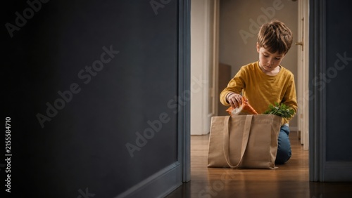 Young boy helps with household chores by unpacking groceries from a reusable canvas tote bag in a sunlit home doorway