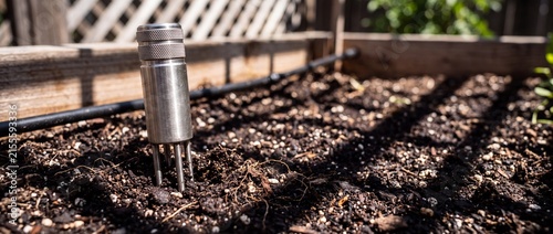 Metal soil sensor probe inserted into dark fertile earth of garden planter box for agricultural moisture monitoring and analysis.