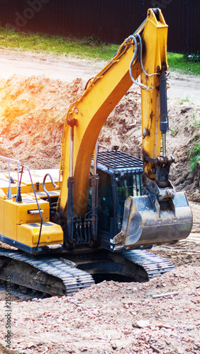 Yellow heavy duty excavator earthmoving machine parked on construction site with dirt piles and green grass under bright sun light lens flare. Vertical photo