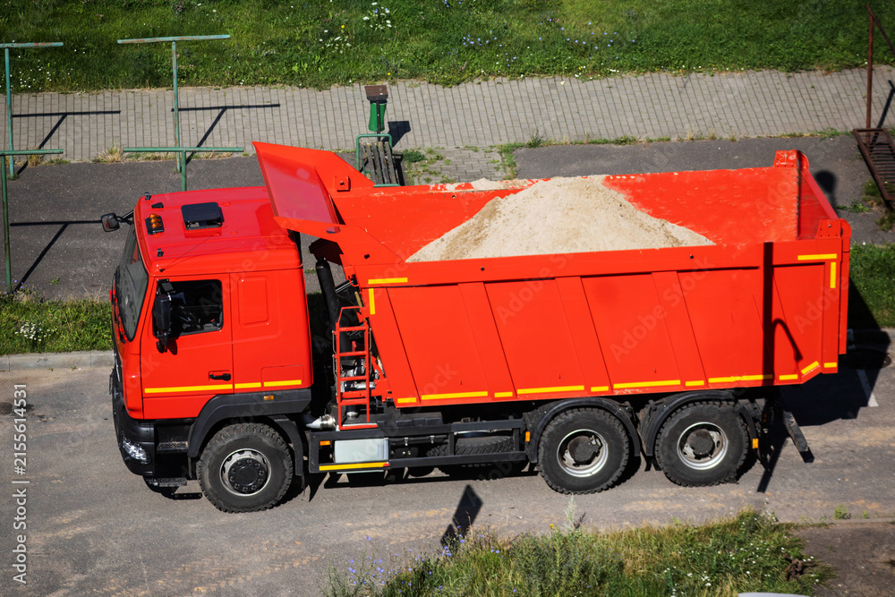 Fototapeta premium Bright red dump truck with open bed loaded with large pile of sand, parked on asphalt road, high angle view captures heavy construction vehicle beside paved sidewalk and green grass.