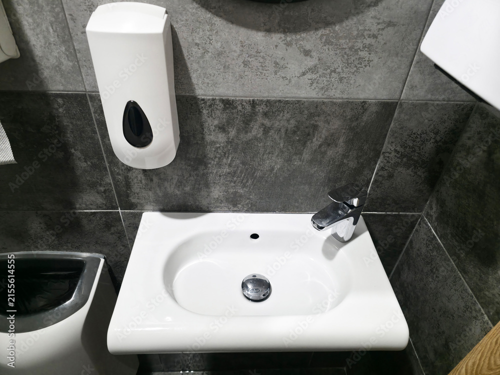 Fototapeta premium Modern public restroom interior white ceramic sink, chrome faucet, wall mounted soap dispenser against dark grey tiled wall for hygiene handwashing.
