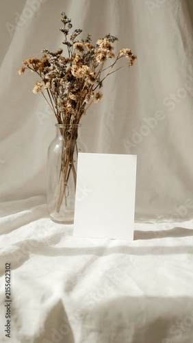 Blank white card mockup with dried flowers in a glass vase on a draped linen fabric background, minimalist aesthetic for wedding stationery
