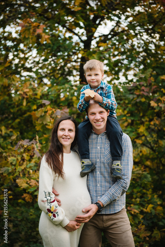 Son sitting on dad's shoulders and hugging mom, happy family portrait in the park in autumn. Pregnant woman and smiling father with child outdoors.