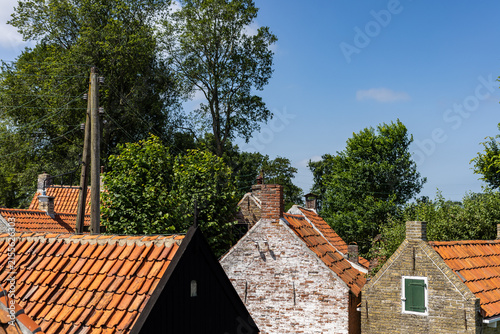 фотографія View over old Dutch rooftops with red clay tiles, brick chimneys, green shutters and trees under a clear blue summer sky