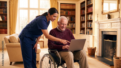 Female caregiver in blue scrubs leaning over senior man in wheelchair pointing at laptop screen in cozy living room