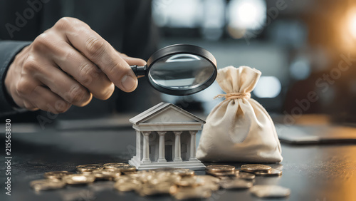 Hand holding a magnifying glass over a miniature bank building beside a tied money bag and scattered coins, financial investigation and anti-money laundering compliance concept