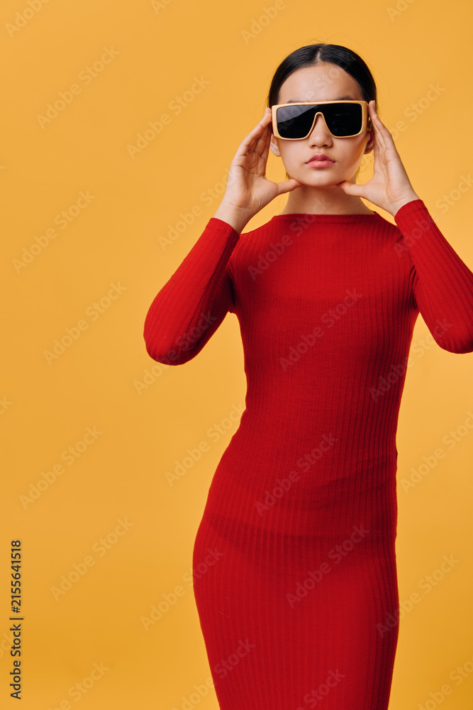 Fototapeta premium Woman in red dress wearing large sunglasses standing against orange background with confident expression and fashion style, studio portrait of female model posing.