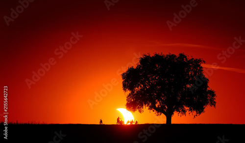Spectacular view of a solar eclipse sunset with the silhouettes of a tree and people on the horizon watching the sun crescent in the deep red vignetted sky. 