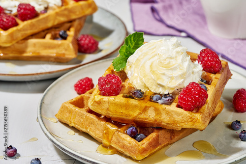 stacked golden brown waffles topped with fresh raspberries and blueberries, whipped cream, and syrup, inviting breakfast dessert option on plates from above close-up.
