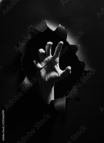 A human hand reaching out through a dark torn paper hole in black and white