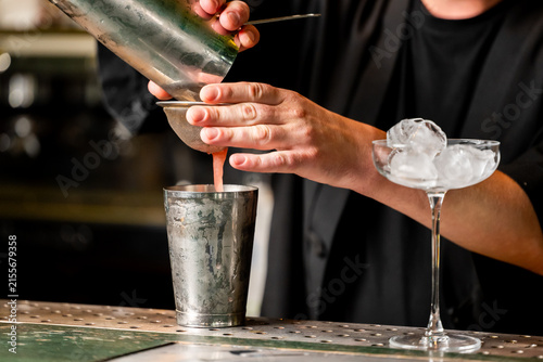 Konstfotografi A bartender straining a cocktail from a metal shaker tin into a metal glass through a fine mesh strainer next to a coupe glass filled with ice on a bar counter