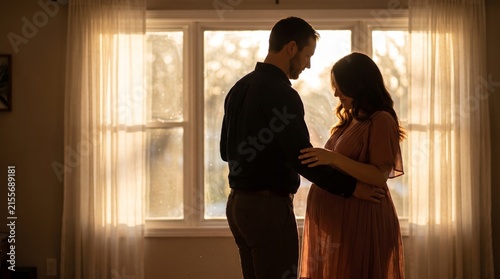 Silhouette of a pregnant couple standing by a window with warm backlighting during golden hour at home