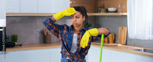 Fotografie Worried Black Female Aged 20s Expression While Cleaning Kitchen With Mop, Wearing Yellow Gloves, Feeling Overwhelmed At Home