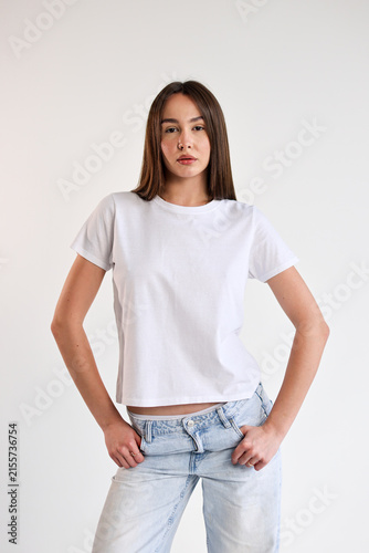 Confident young woman standing against plain white background wearing white t shirt and light jeans with relaxed pose, straight hair, neutral expression, minimal studio portrait with clean copy space