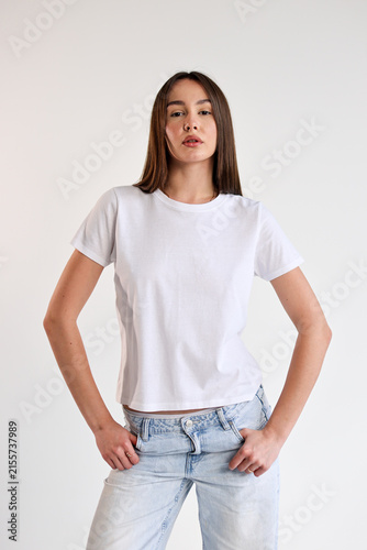Young woman standing confidently in white t shirt and light denim jeans against plain white background with hands on hips, relaxed posture, clean studio portrait, minimal fashion look