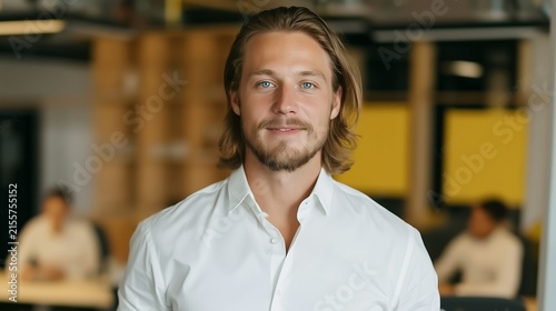 cheerful young businessman looking at camera in office setting with team members working behind him illustrating modern career and corporate portrait concept
