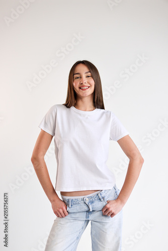 Smiling young woman in blank white t shirt and light jeans standing against clean white background with hands on hips, relaxed confident posture, casual fashion portrait with ample copy space