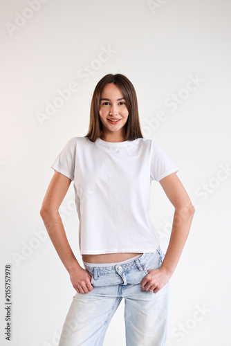Confident smiling young woman in plain white t shirt and light blue jeans posing with hands on hips against clean white background, casual fashion portrait with minimal modern style