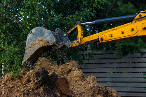 Fotografie Close up of yellow excavator bucket dumping dirt soil on pile near backyard wooden board fence