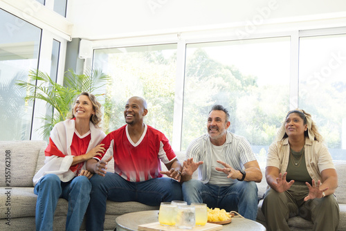 Diverse friends cheering on gray sofa at home by coffee table, two wearing red sports tops