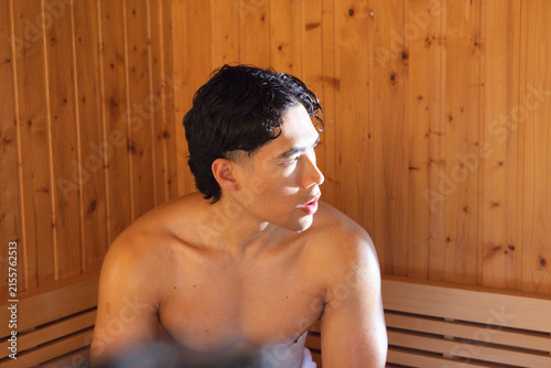 Canvas Print Asian man sitting shirtless on sauna bench, looking right as warm light highligh