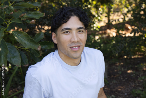 Asian man sitting in garden beside broad green leaves on left wearing light crew-neck T-shirt