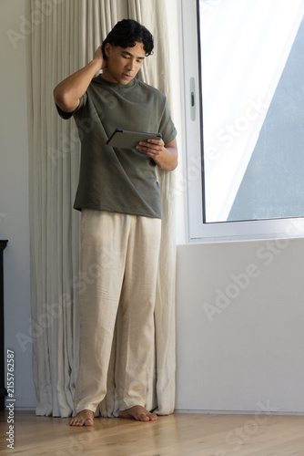 Foto Man standing barefoot at home wearing olive T-shirt and linen pants, holding tab