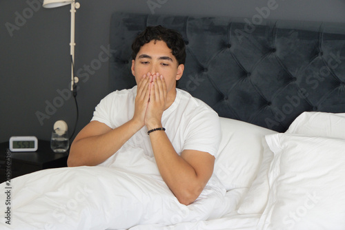 Konstfotografi Asian man sitting up in bed in bedroom wearing white T-shirt covering mouth near