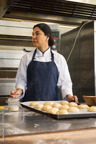 фотографія Woman chef in white jacket, dark apron preparing dough rolls on stainless-steel