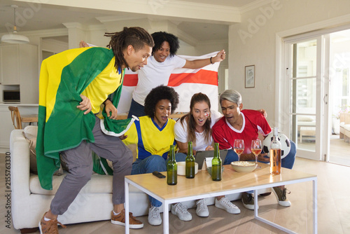 Diverse friends in jerseys cheering and watching tablet on sofa with beer bottles and soccer ball