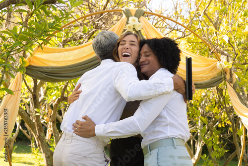 Diverse friends hugging under yellow drape arch in garden in dressy wear holding tablet