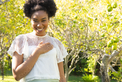 African American woman standing, smiling hand on chest in orchard wearing white-lace and gold-hoops