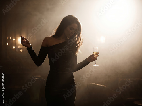 Elegant woman in a velvet dress holding a glass in a moody hazy lounge, symbolizing premium nightlife and introspective solitude.