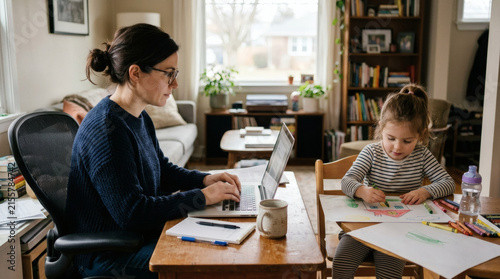 A focused mother is diligently working on her laptop at a home desk while her young daughter peacefully draws at a small table beside her, balancing remote work and crucial childcare responsibilities.