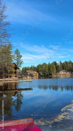 Peaceful lakeside homes beside still water with bright red boat edge, narrow dock, mirrored reflections, forest shoreline, clear blue sky, soft clouds, and calm rural vacation setting
