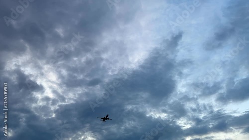 Small aircraft silhouette crossing beneath layered clouds in wide blue gray sky with bright openings, moody diffused daylight, strong negative space, and expansive atmospheric backdrop