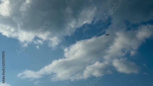 Small airplane silhouette crossing bright blue sky beneath large white clouds with open airy space, calm daylight atmosphere, expansive upward view, soft contrast, travel and aviation concept