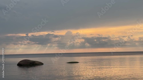 Calm sunset seascape with glowing clouds over reflective water, dark foreground rocks, low horizon, soft golden light, spacious coastal mood and tranquil natural atmosphere