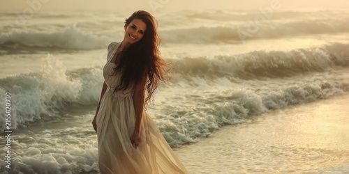 Radiant young woman, long dark wavy, flowing hair standing, posing gracefully on the sand beach, ocean ,sea waves shoreline while wearing a light white dress during a warm golden evening sunset glow