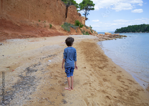 Little boy standing on a unique golden sand beach with reddish clay cliffs on Vrgada island, Croati