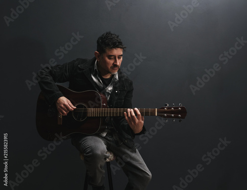 Foto Músico hispano con chaqueta negra tocando una guitarra acústica con pasión, sentado en estudio con fondo oscuro