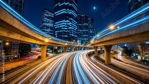 Expansive urban highway system at night with striking blue light trails, towering skyscrapers, crescent moon, city lights, modern infrastructure, dynamic traffic, and futuristic metropolitan atmosph