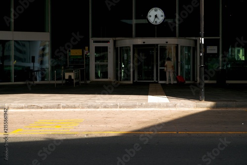 Woman with red luggage entering modern train station terminal at taxi zone under clock in harsh sunlight