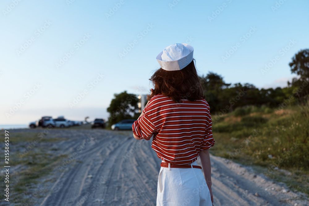 Obraz premium Back view of a woman in striped shirt and skirt along a dirt road by the beach, capturing lifestyle, outdoor travel and casual summer fashion.