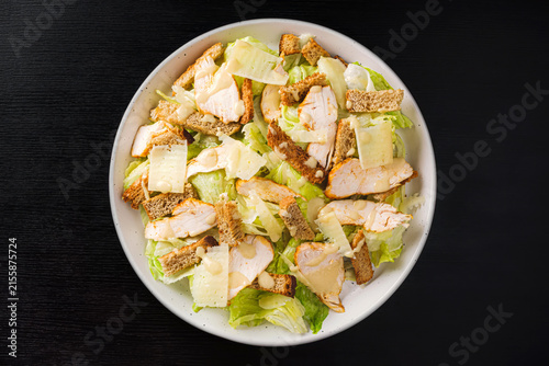 Top view of Caesar salad with grilled chicken, croutons, parmesan shavings and dressing on romaine lettuce, white plate on black wood table, copy space
