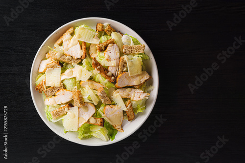 Top view of Caesar salad with grilled chicken, croutons, parmesan shavings and dressing on romaine lettuce, white plate on black wood table, copy space