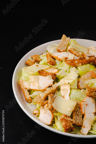 Top view of Caesar salad with grilled chicken, croutons, parmesan shavings and dressing on romaine lettuce, white plate on black wood table, copy space