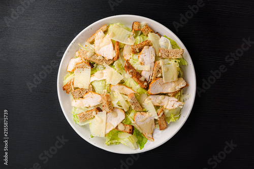 Top view of Caesar salad with grilled chicken, croutons, parmesan shavings and dressing on romaine lettuce, white plate on black wood table, copy space